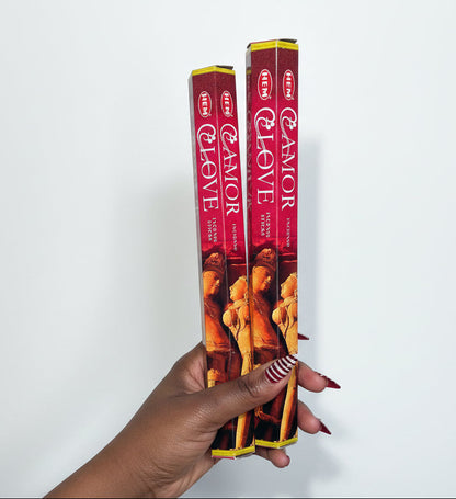 Two boxes of HEM Amor y Love incense sticks held by a hand on a white surface.