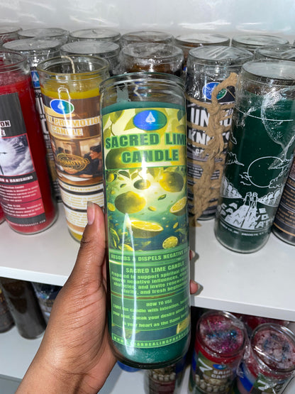 A hand holding a tall glass jar candle labeled 'Sacred Lime Candle' with green and yellow graphics, set against a background of other candles on a white shelf.